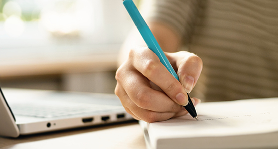 Bildausschnitt einer Hand mit Stift, schreibt in Notizbuch, Laptop neben Notizbuch